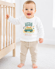 A smiling baby boy wearing a white sweatshirt with a graphic design and the name "Olivier" printed on it, standing in front of a wooden crib.