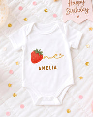 A white onesie with a red strawberry and the name "Amelia" printed on it, surrounded by confetti and a birthday card.