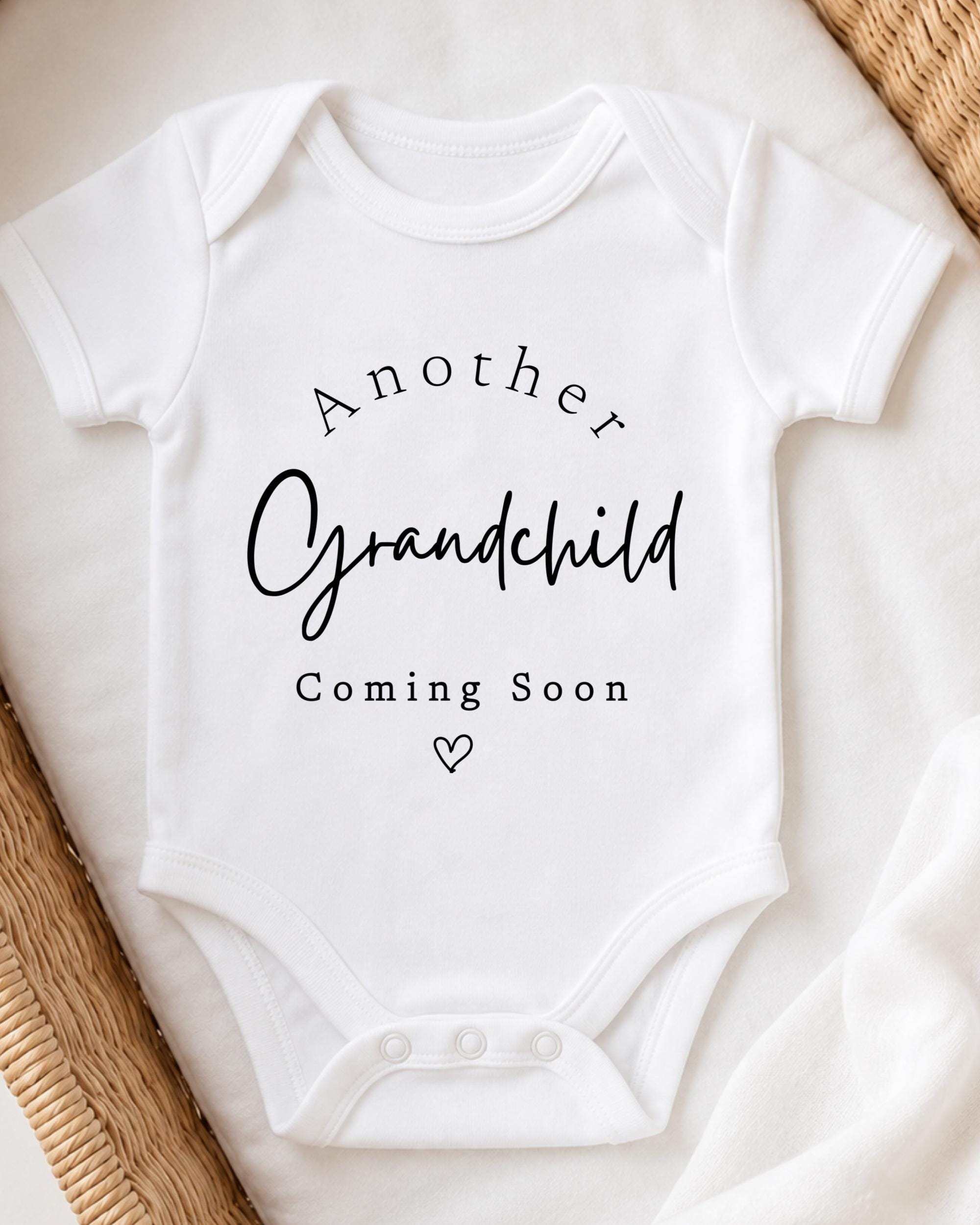A white baby onesie with the text "Another Grandchild Coming Soon" printed on it.