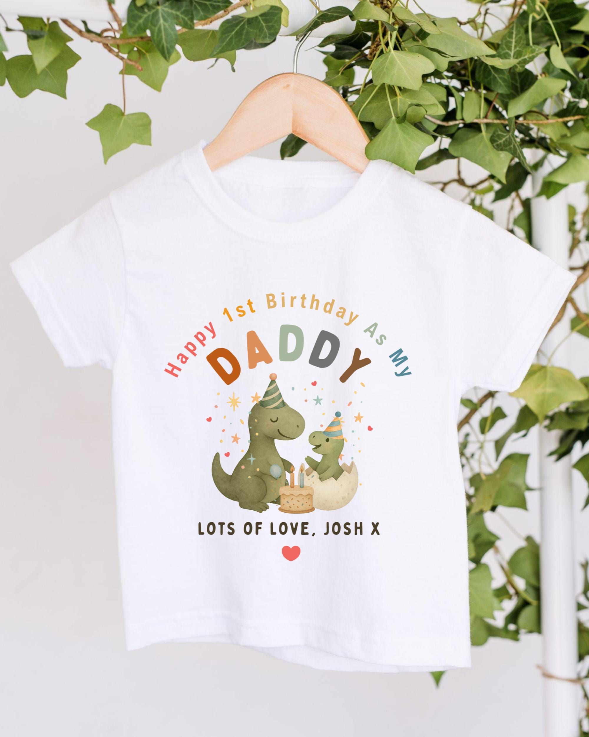A white t-shirt with a colorful graphic design featuring a dinosaur and a baby dinosaur, along with the text "Happy 1st Birthday As My Daddy Lots of Love Josh X".