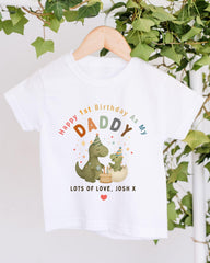 A white t-shirt with a colorful graphic design featuring a dinosaur and a baby dinosaur, along with the text "Happy 1st Birthday As My Daddy Lots of Love Josh X".
