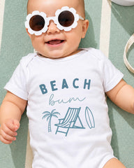 A baby wearing a white onesie with the text "BEACH butt" and a beach-themed graphic, smiling and laying on a green and white striped surface.