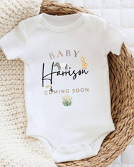 A white baby onesie with the text "BABY Harrison" and "COMING SOON" printed on it, along with an illustration of a giraffe and a zebra.