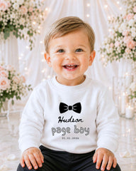A smiling baby boy wearing a white sweater with a black bow tie and the text "Hudson page boy" printed on it.