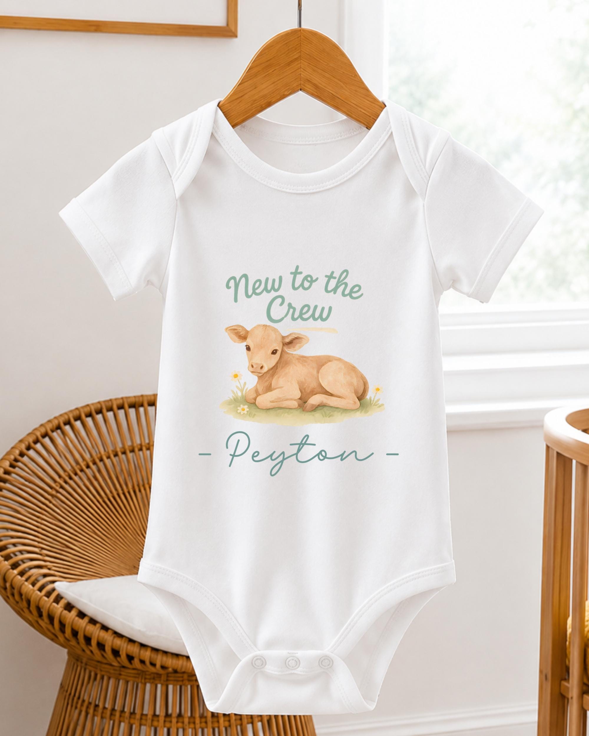 A white baby onesie with a printed image of a calf and the text "New to the Crew - Peyton" printed on it, hanging on a wooden hanger.