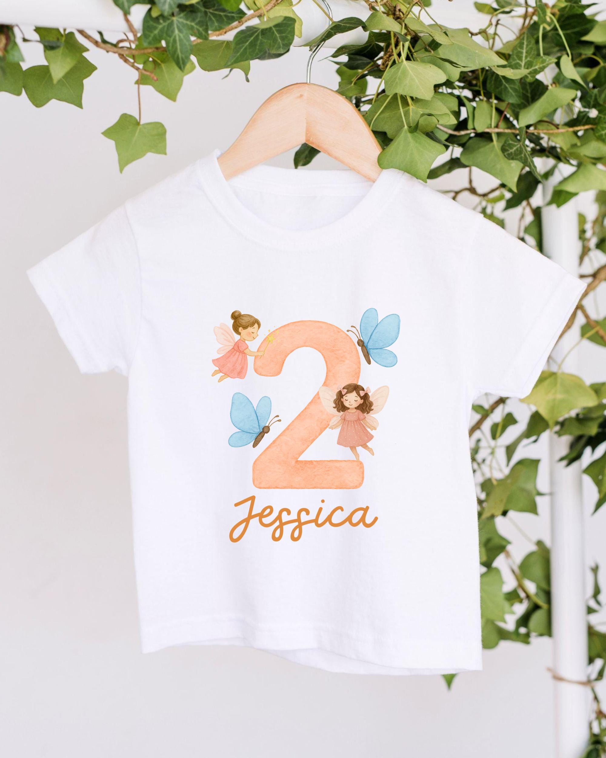 A white t-shirt with a personalized design hanging on a wooden hanger, featuring a large number "2" with two small fairy characters and the name " Jessica" written below.