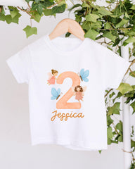 A white t-shirt with a personalized design hanging on a wooden hanger, featuring a large number "2" with two small fairy characters and the name " Jessica" written below.