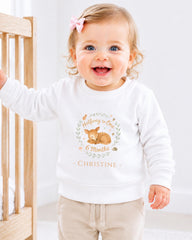 A smiling baby girl wearing a white sweatshirt with a deer graphic and the text "Halfway to One 6 Months - Christine" printed on it.