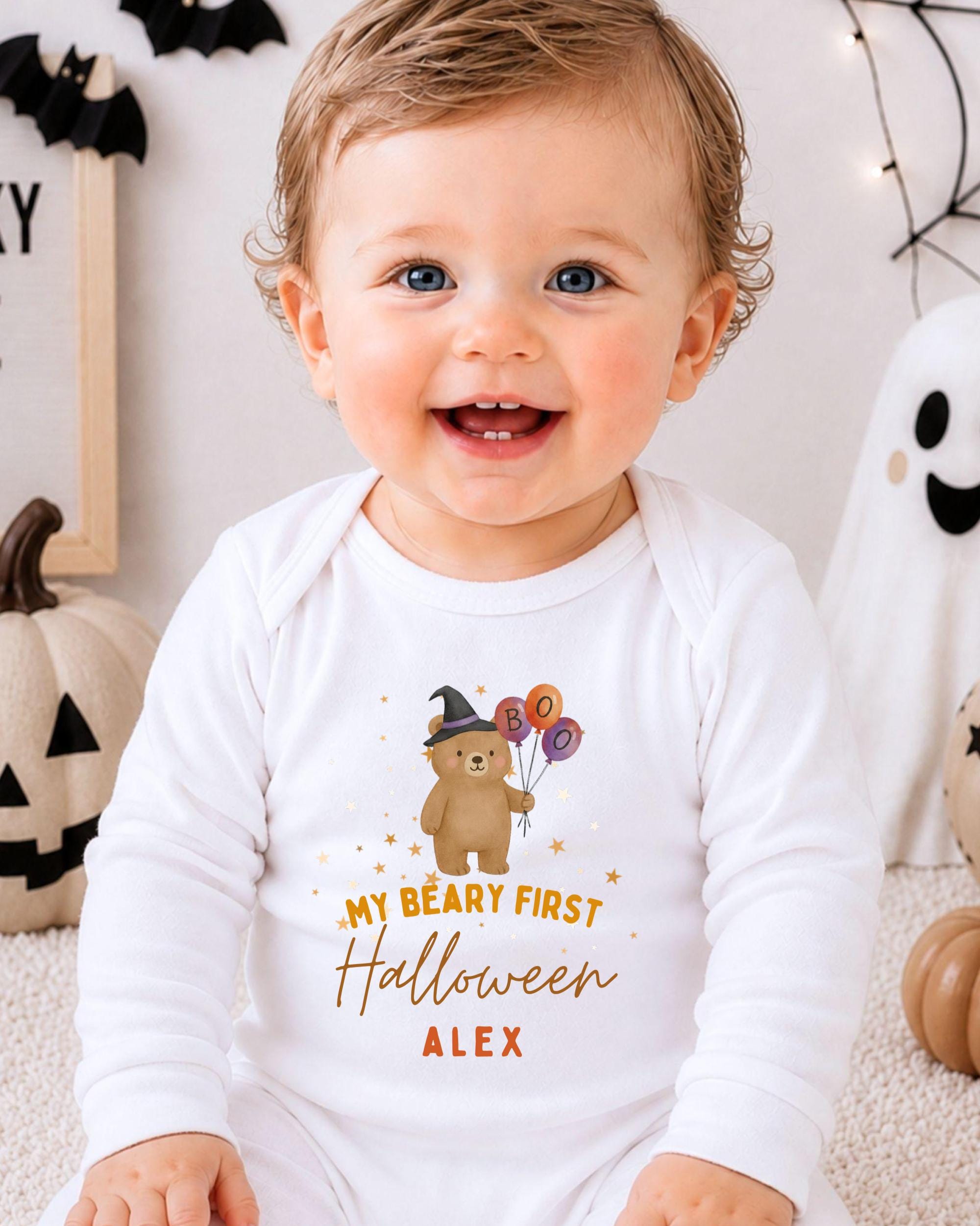 A smiling baby wearing a white onesie with a Halloween-themed design, including a teddy bear and balloons.