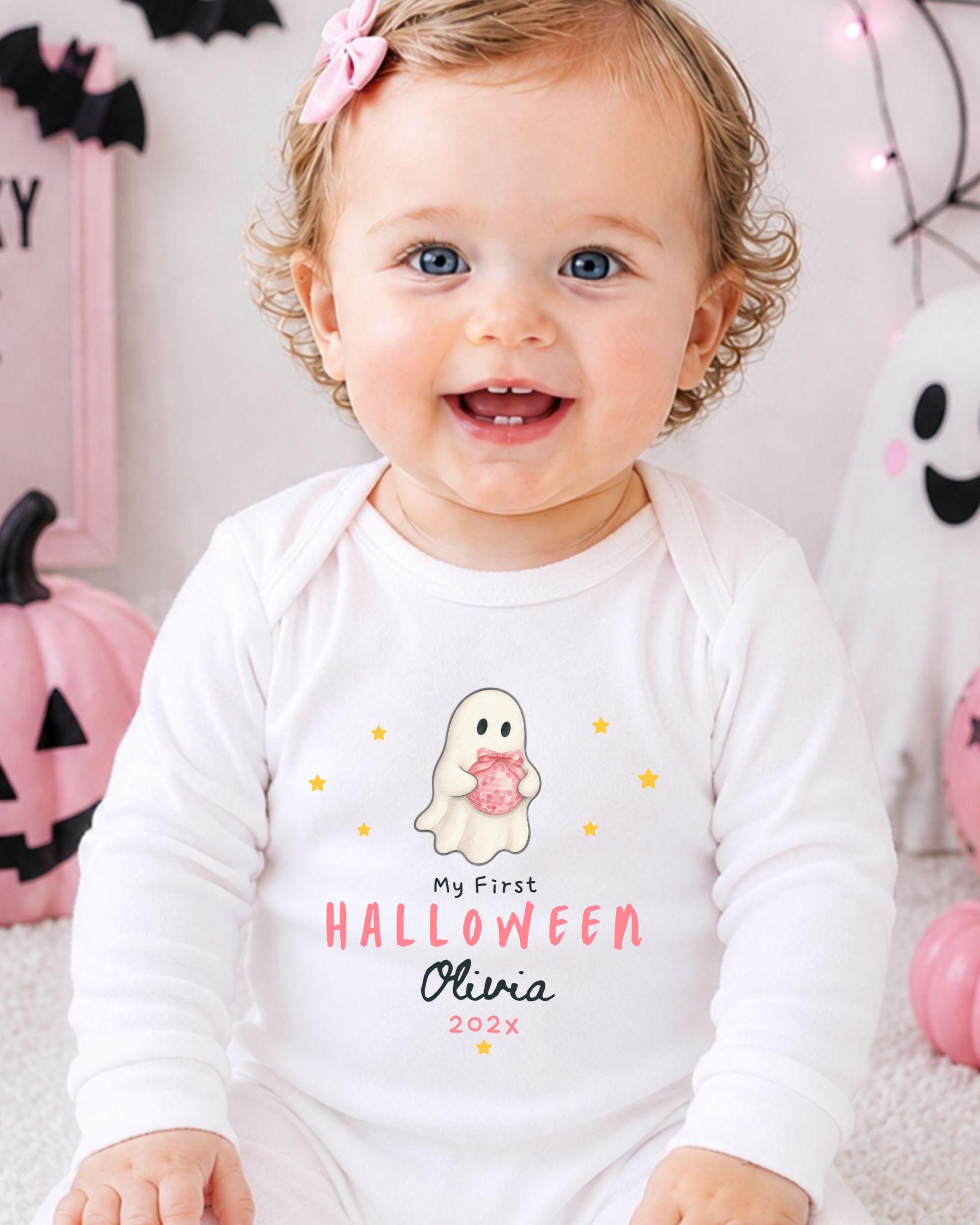 A smiling baby girl wearing a white onesie with a ghost and text, sitting in front of Halloween decorations.