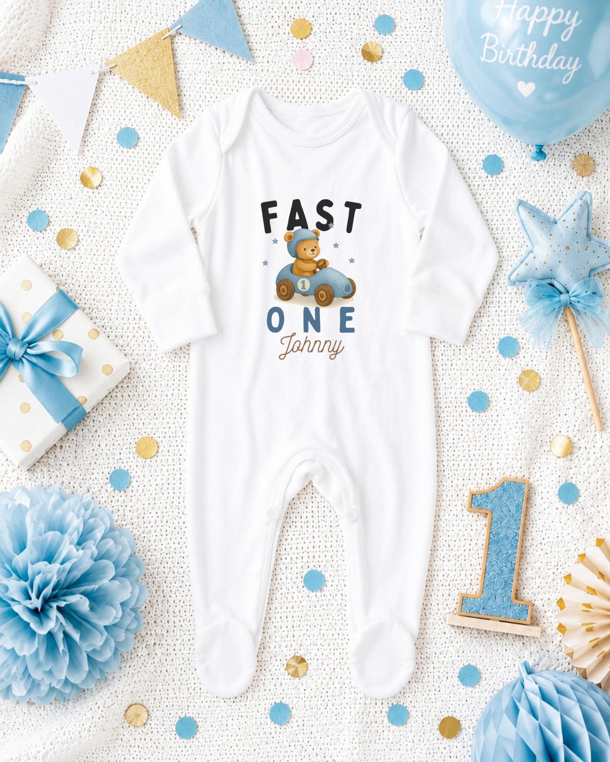 A white onesie with the text "FAST ONE Johnny" printed on it, surrounded by various baby-themed items such as a blue ribbon, a blue star, a blue and gold gift box, and a blue and gold pom pom.