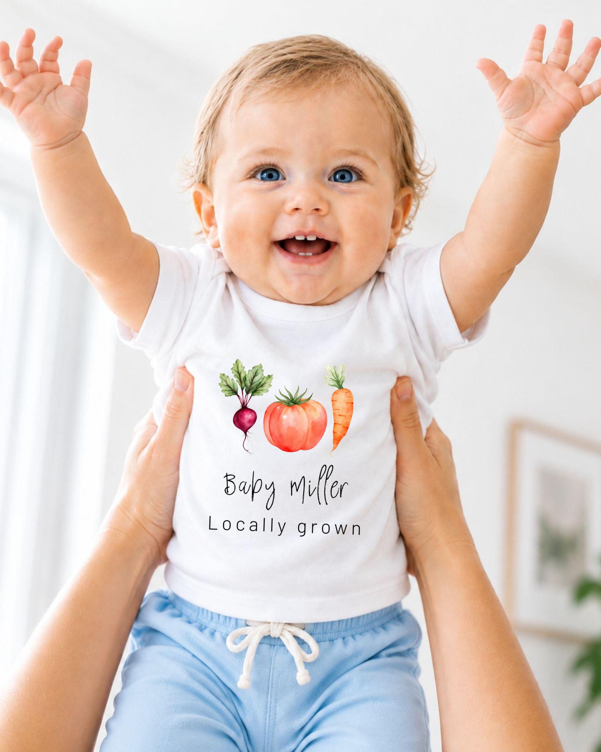 A smiling baby wearing a white t-shirt with a vegetable design and the name "Baby Muller" printed on it, being held up by someone's arms.
