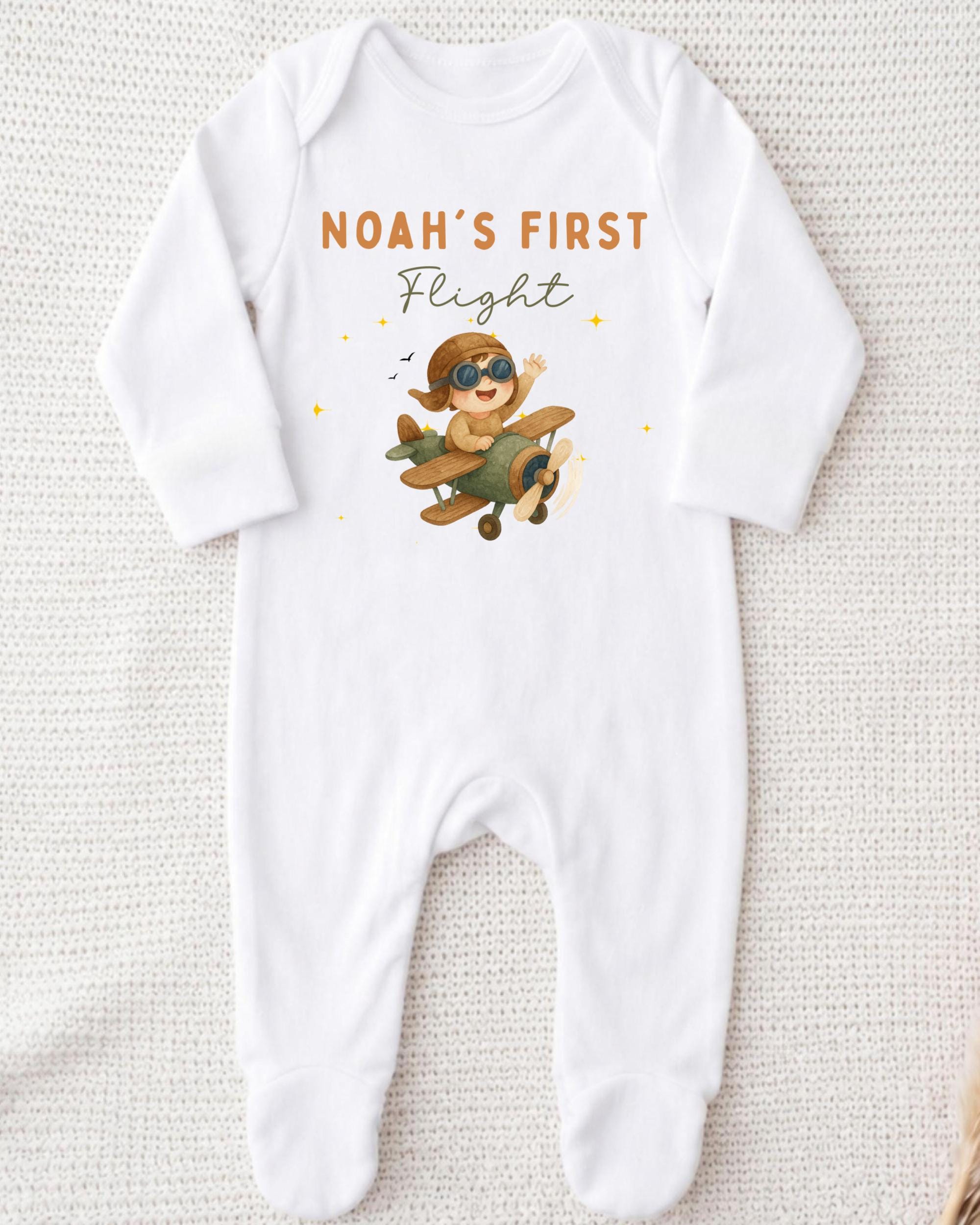 A white onesie with a cartoon image of a baby wearing a pilot's hat and flying an airplane, along with the text "Noah's first flight".