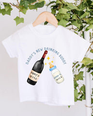 A white t-shirt with a graphic design featuring a bottle of wine and a baby bottle, along with the text "DADY'S NEW DRINKING BUDDY" and "JOSH'S MILK" printed on it.