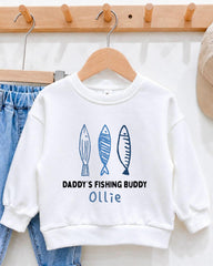 A white sweatshirt with a custom design featuring three fish illustrations and the text "DADDY'S FISHING BUDDY OLLIE" printed on it.