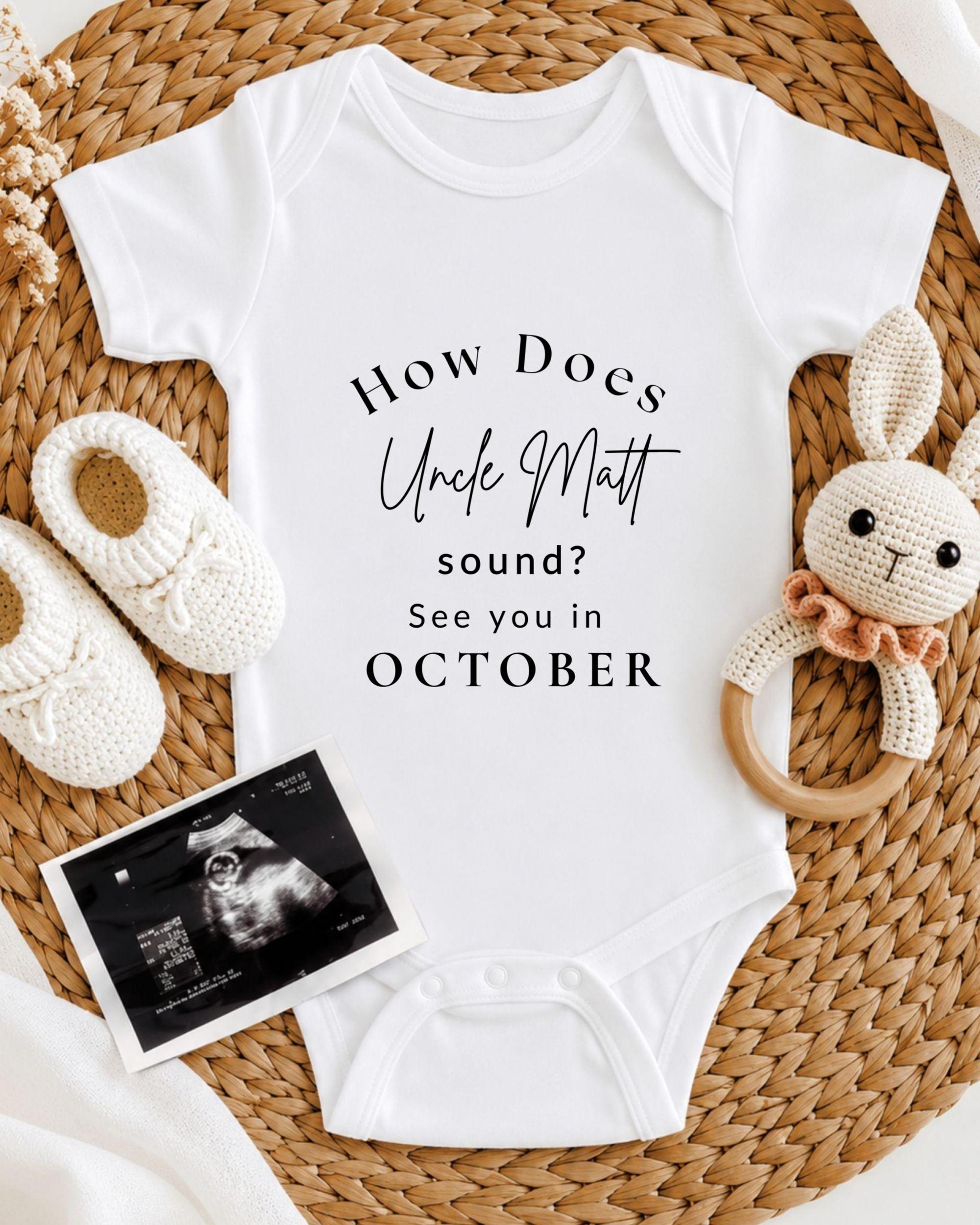 A white baby onesie with text, a crocheted bunny toy, and a baby bootie are arranged on a woven mat.