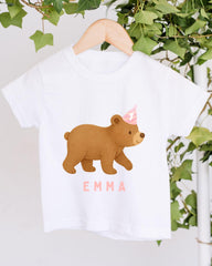 A white t-shirt with a cartoon bear wearing a party hat and the name "Emma" printed on it, hanging on a wooden hanger.