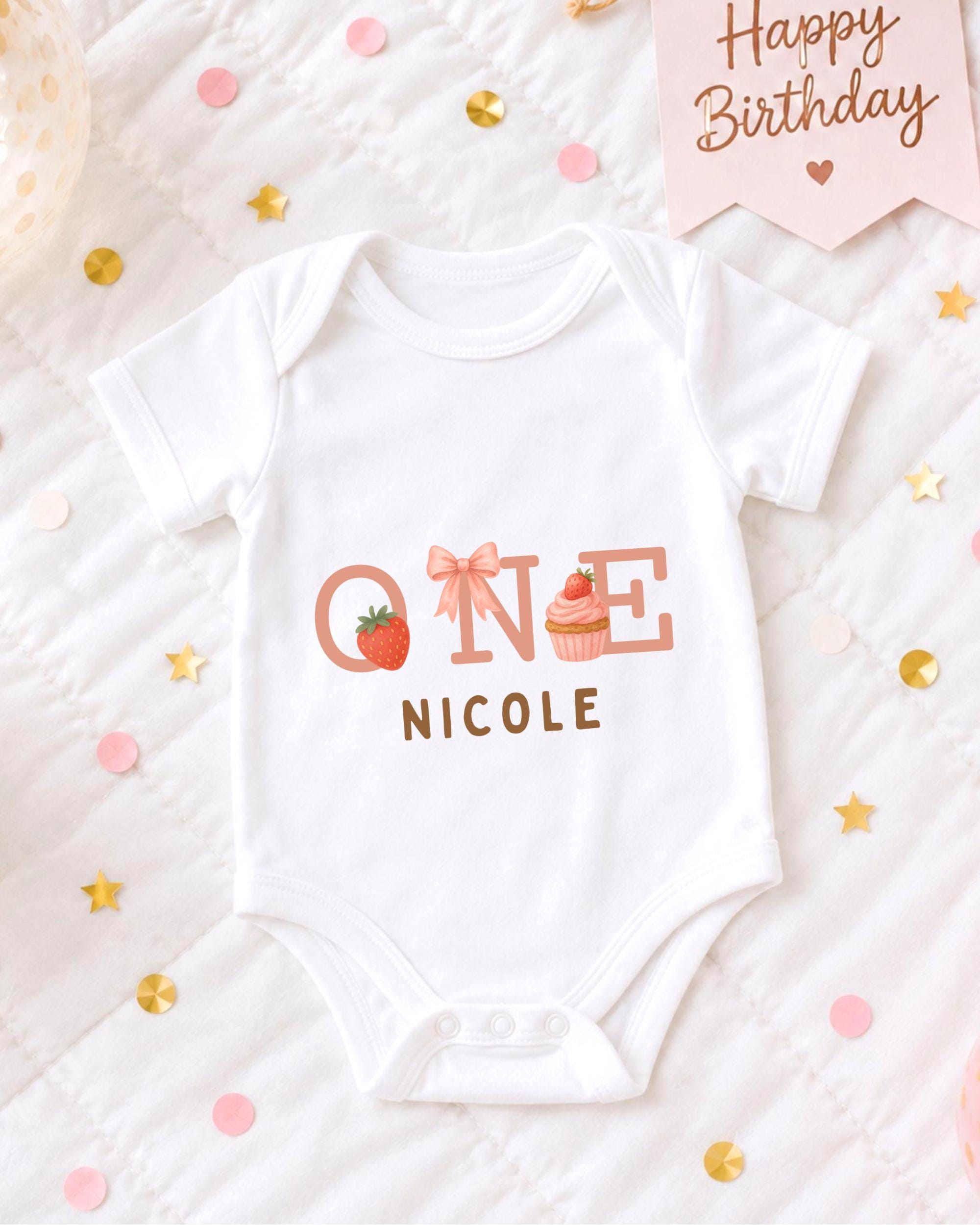 A white onesie with the text "ONE NICOLE" printed on it, along with illustrations of a strawberry and a cupcake, is displayed on a bed with confetti and a "Happy Birthday" sign.