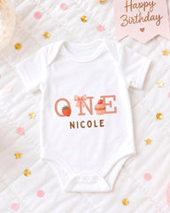 A white onesie with the text "ONE NICOLE" printed on it, along with illustrations of a strawberry and a cupcake, is displayed on a bed with confetti and a "Happy Birthday" sign.