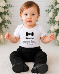 A smiling baby boy wearing a white shirt with a bow tie and black pants sits on a white carpet, with a floral background behind him.