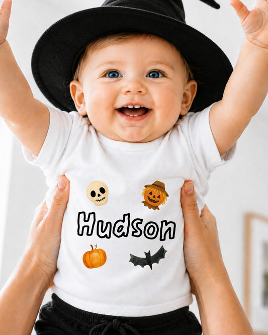 A smiling baby wearing a white t-shirt with Halloween-themed designs and the name "Hudsons" printed on it.