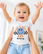A smiling baby wearing a white t-shirt with a graphic design and text, being held up by someone's arms.