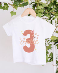 A white t-shirt with the number 3 and butterflies printed on it, hanging on a wooden hanger.