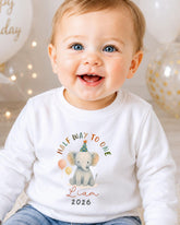 A smiling baby boy wearing a white shirt with a graphic of an elephant and the text "Half way to one Liam 2026" printed on it.