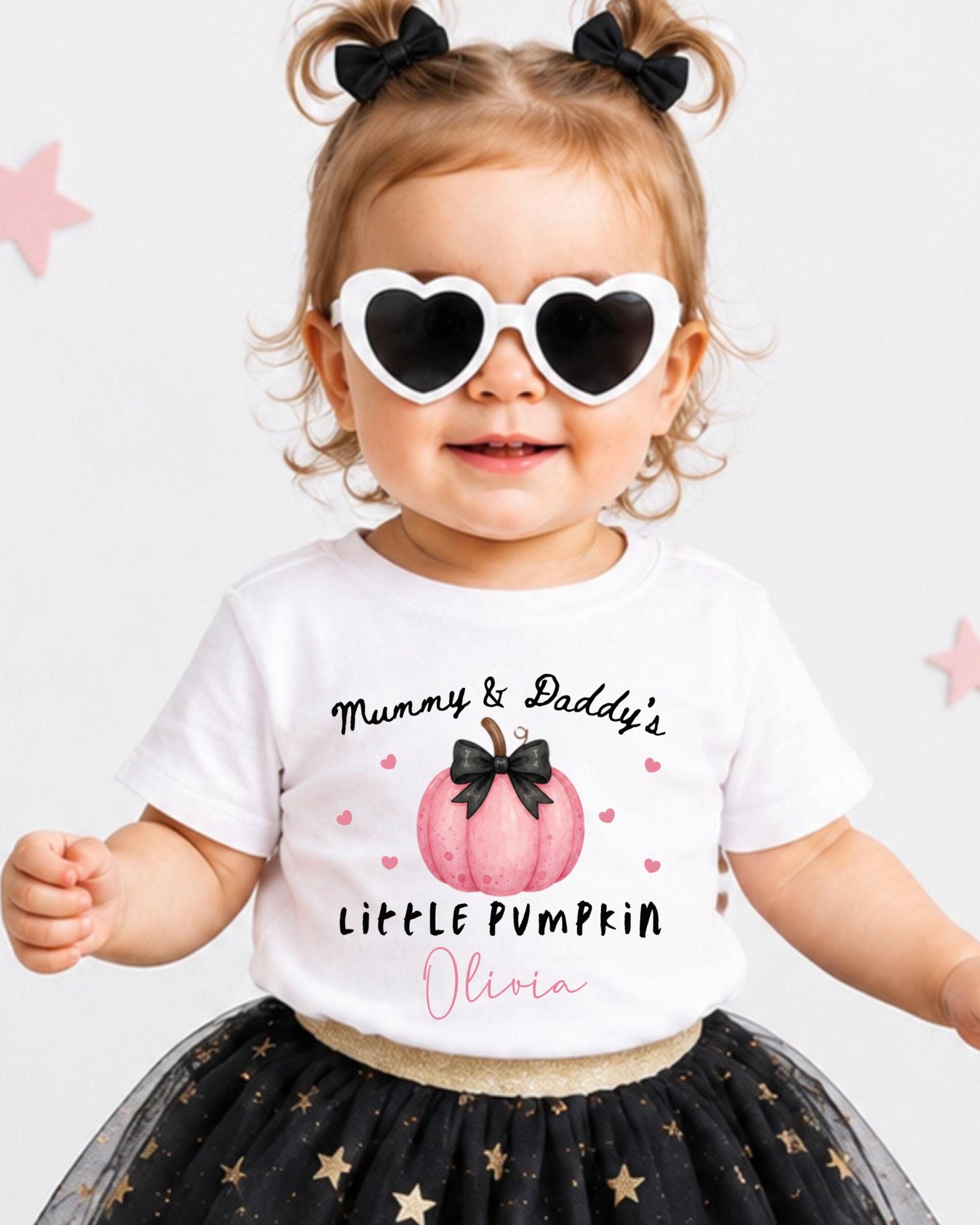 A young girl wearing a white t-shirt with a pumpkin graphic and the text "Mummy & Daddy's LITTLE PUMPKIN Olivia" is smiling and wearing heart-shaped sunglasses.