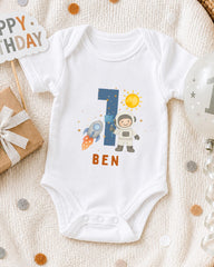 A white onesie with a space theme design, featuring a rocket ship, a person in a spacesuit, and the number 1. The word "BEN" is written below the image.