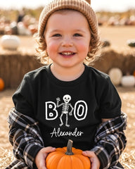 A young child wearing a black t-shirt with a skeleton design and the name "Alexander" printed on it, holding an orange pumpkin.