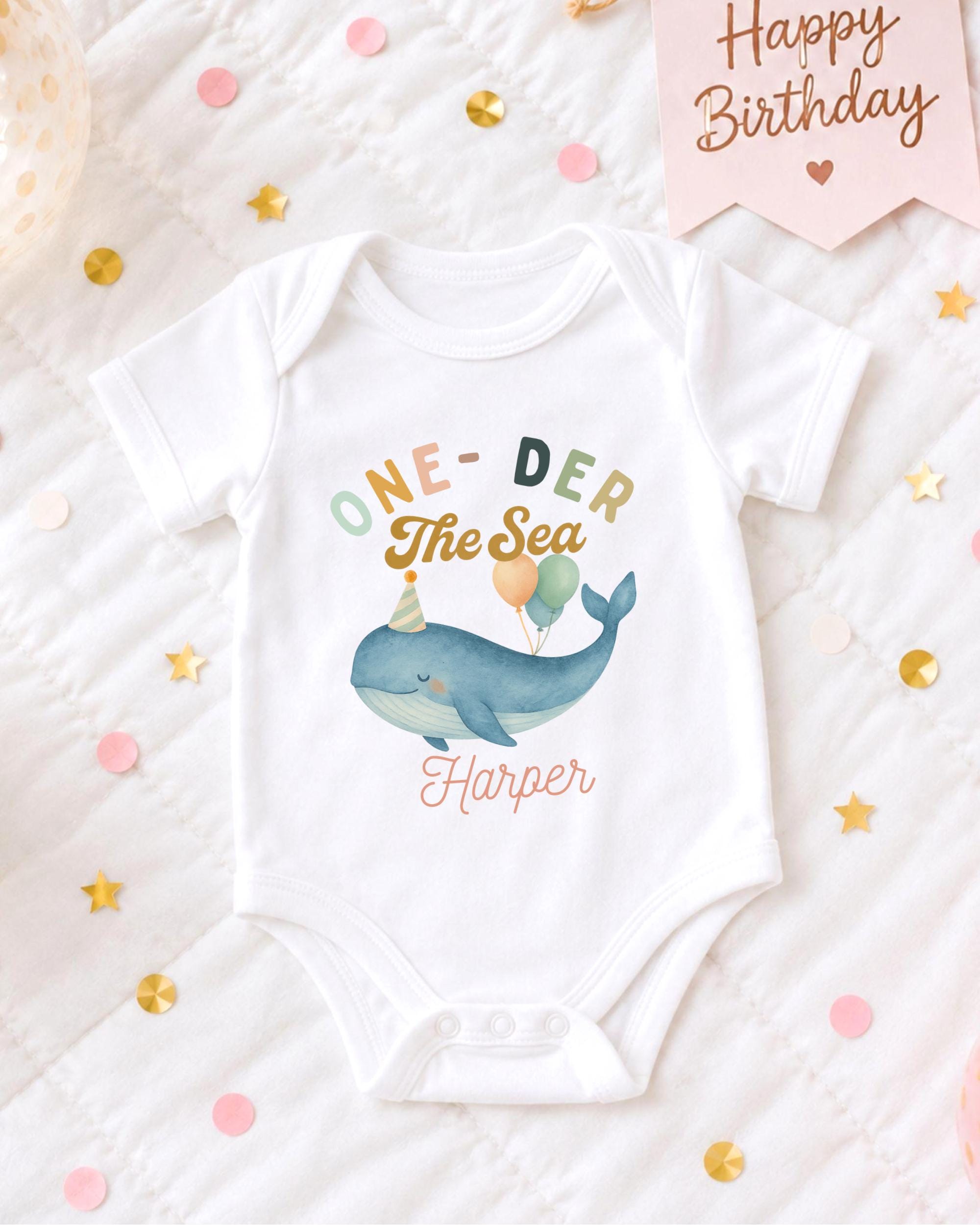 A white onesie with a whale design and the text "One-DER The Sea Harper" printed on it, along with confetti and a birthday card.