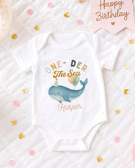 A white onesie with a whale design and the text "One-DER The Sea Harper" printed on it, along with confetti and a birthday card.