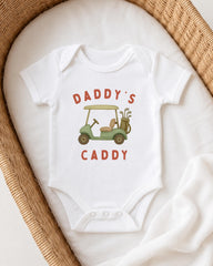 A white onesie with a golf cart design and the text "DADDY'S CADDY" printed on it, laid out on a white cloth.
