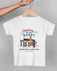 A white t-shirt with a graphic design depicting a race scene, including a finish line, runners, and a clock. The text on the t-shirt reads "Cheering for my DADDY" and "Marathon Name Here" along with "CUSTOM DATE".