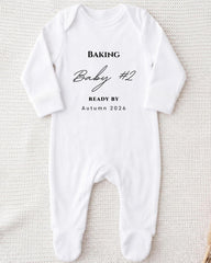 A white onesie with the text "BAKING Baby #2 READY BY Autumn 2026" printed on it.