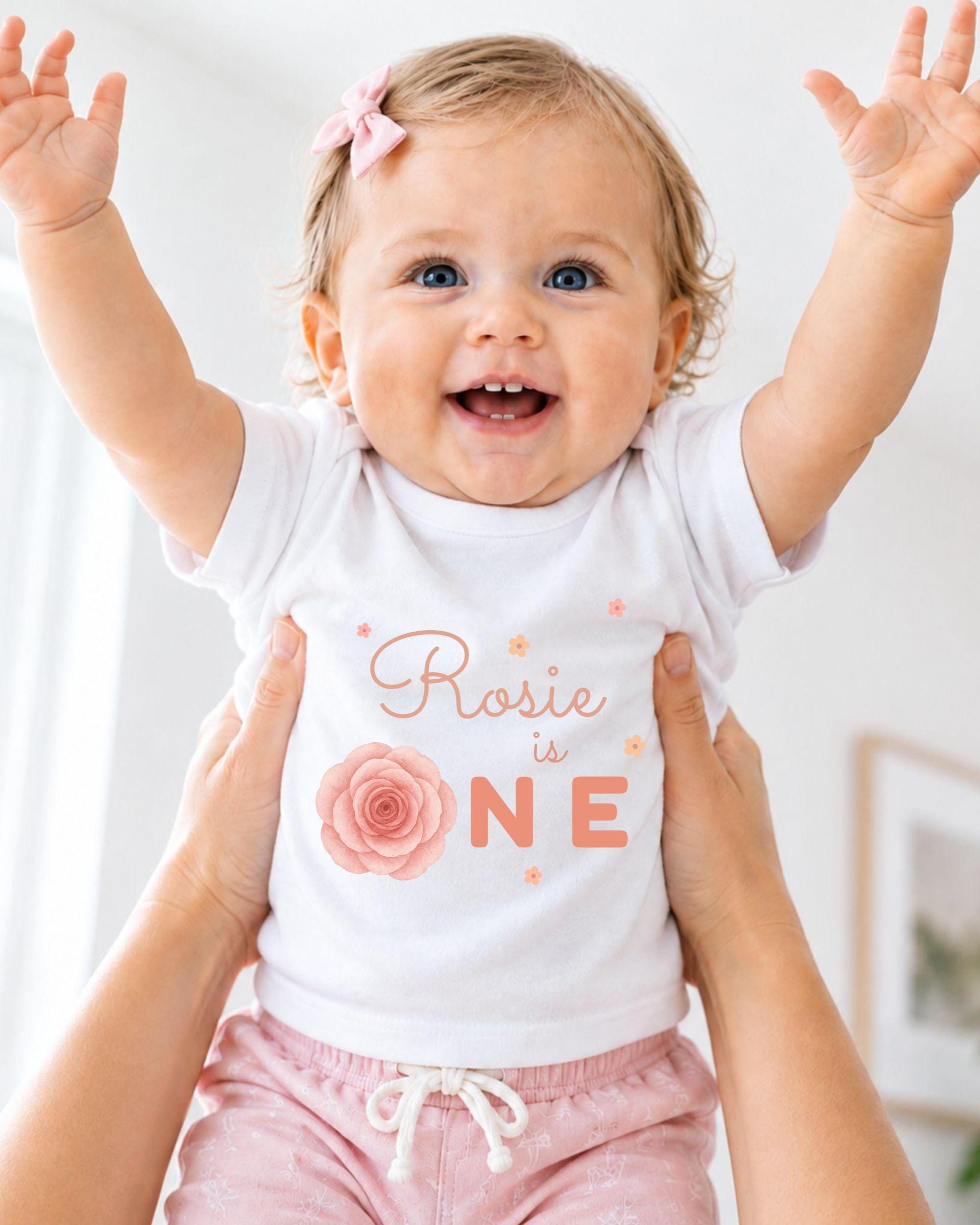 A smiling baby girl wearing a white shirt with a rose design and the text "Rosa is one" is being held up by someone's arms.