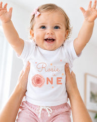 A smiling baby girl wearing a white shirt with a rose design and the text "Rosa is one" is being held up by someone's arms.