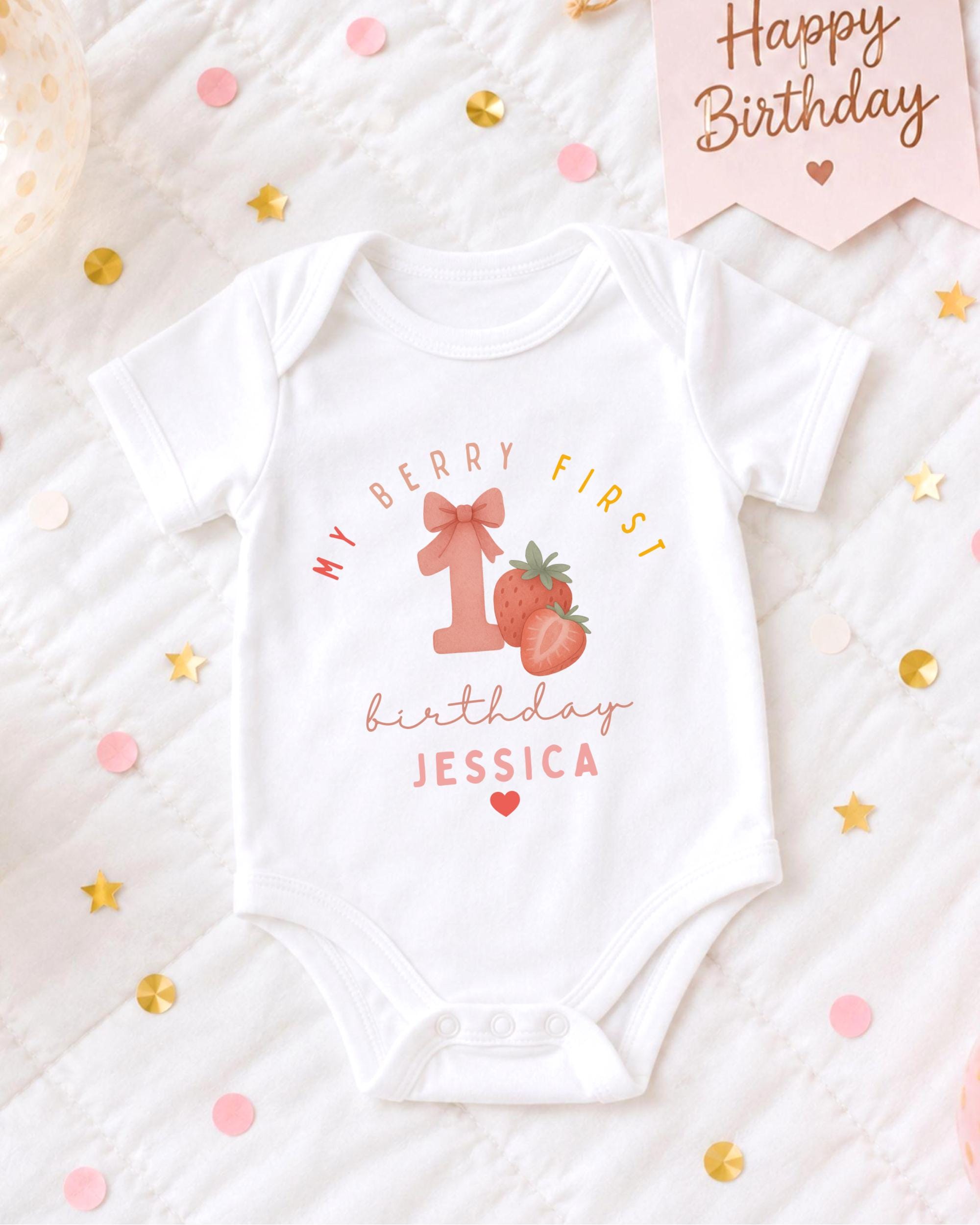 A white onesie with a personalized message and a strawberry graphic, placed on a bed with confetti and a birthday card.