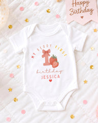 A white onesie with a personalized message and a strawberry graphic, placed on a bed with confetti and a birthday card.