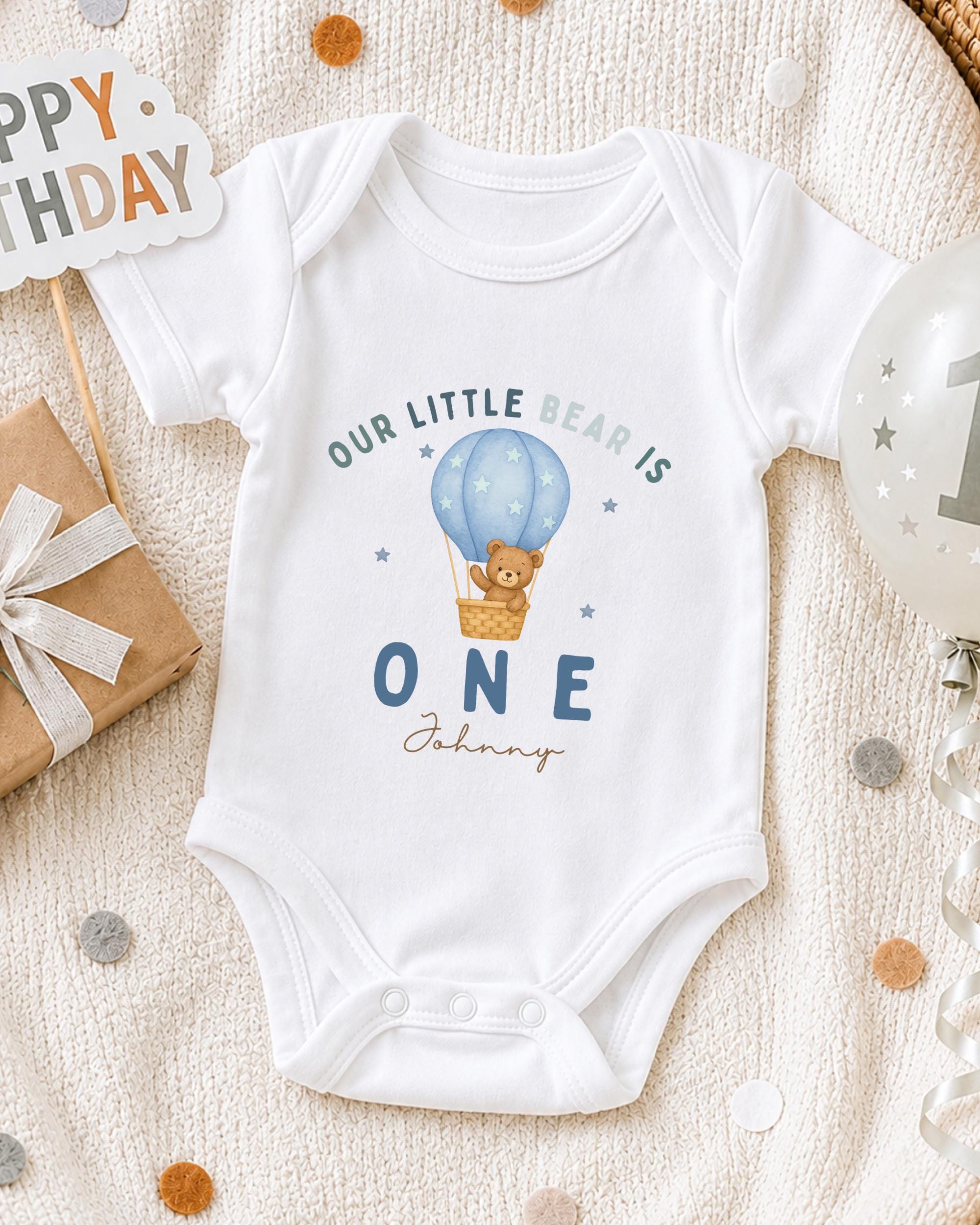A white onesie with a teddy bear and a hot air balloon graphic, along with a "Happy Birthday" sign and a birthday balloon, all arranged on a white blanket.