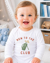A smiling baby wearing a white onesie with the text "NEW TO THE CLUB" printed on it, sitting in a room with a teddy bear and bookshelf in the background.