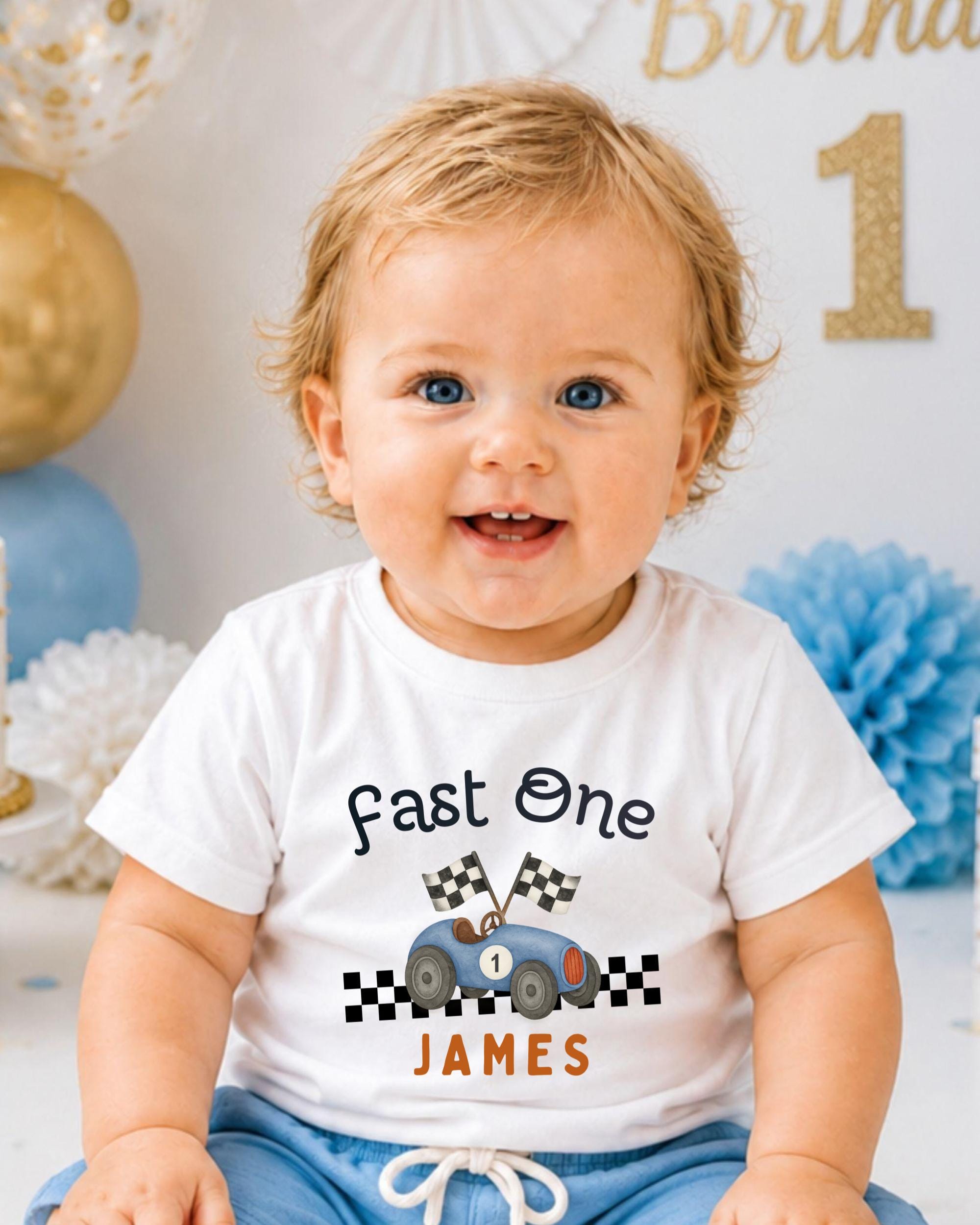 A smiling baby boy wearing a white t-shirt with a racing car graphic and the text "Fast One James" is sitting in front of a birthday party backdrop.