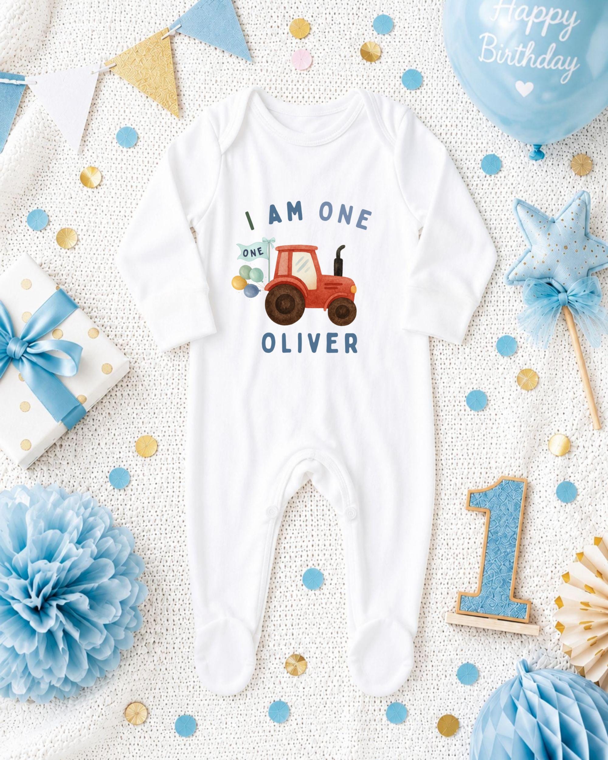 A white onesie with a tractor design, surrounded by birthday decorations such as a blue star, blue and gold confetti, and a blue ribbon.