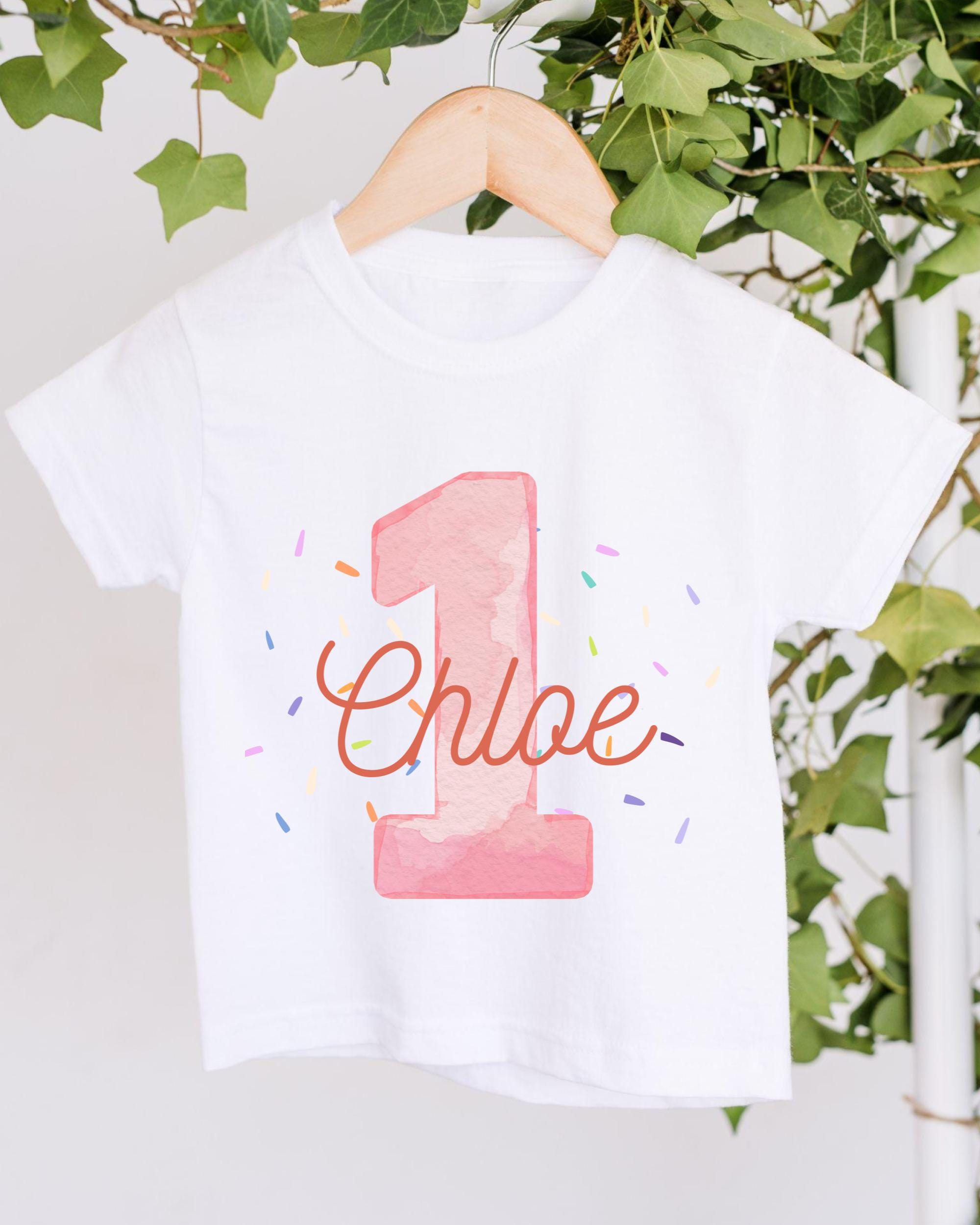 A white t-shirt with a pink number one and the name "Chloe" printed on it, hanging on a wooden hanger.