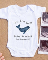 A white baby onesie with a graphic of a blue whale and the text "Sea You Soon Baby Seahell Due December 2027" printed on it, along with a photograph of an ultrasound image of a baby.