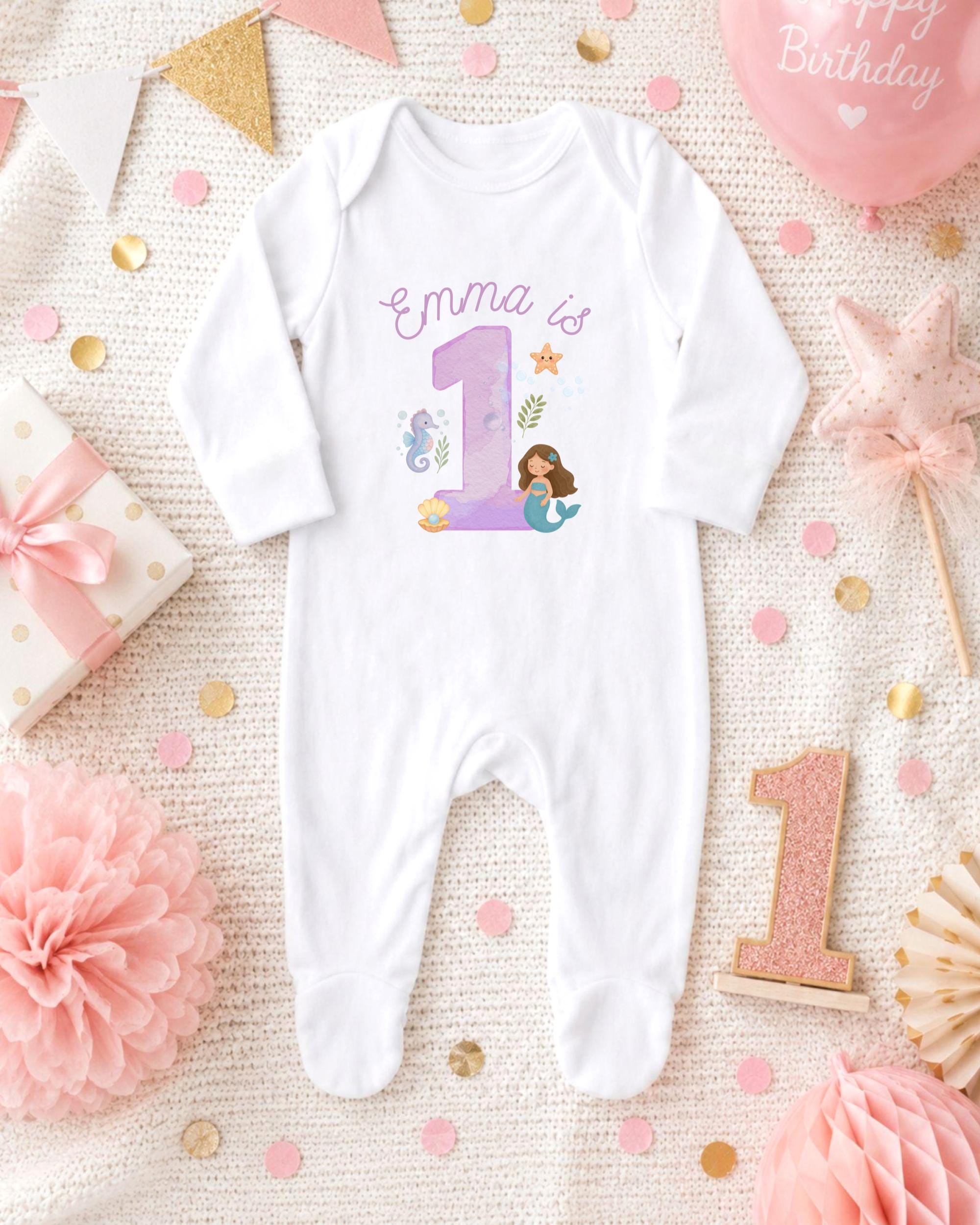 A white onesie with a mermaid design and the number 1, surrounded by pink and gold decorations, including a pink star, confetti, and a pink ribbon.