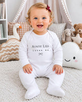 A smiling baby girl wearing a white onesie with the text "Auntie Lisa loves me" sits on a white carpet surrounded by stuffed animals.