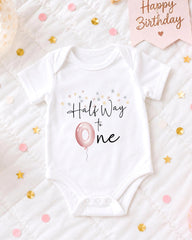 A white onesie with the text "Hals Way to One" printed on it, along with a pink balloon and confetti.