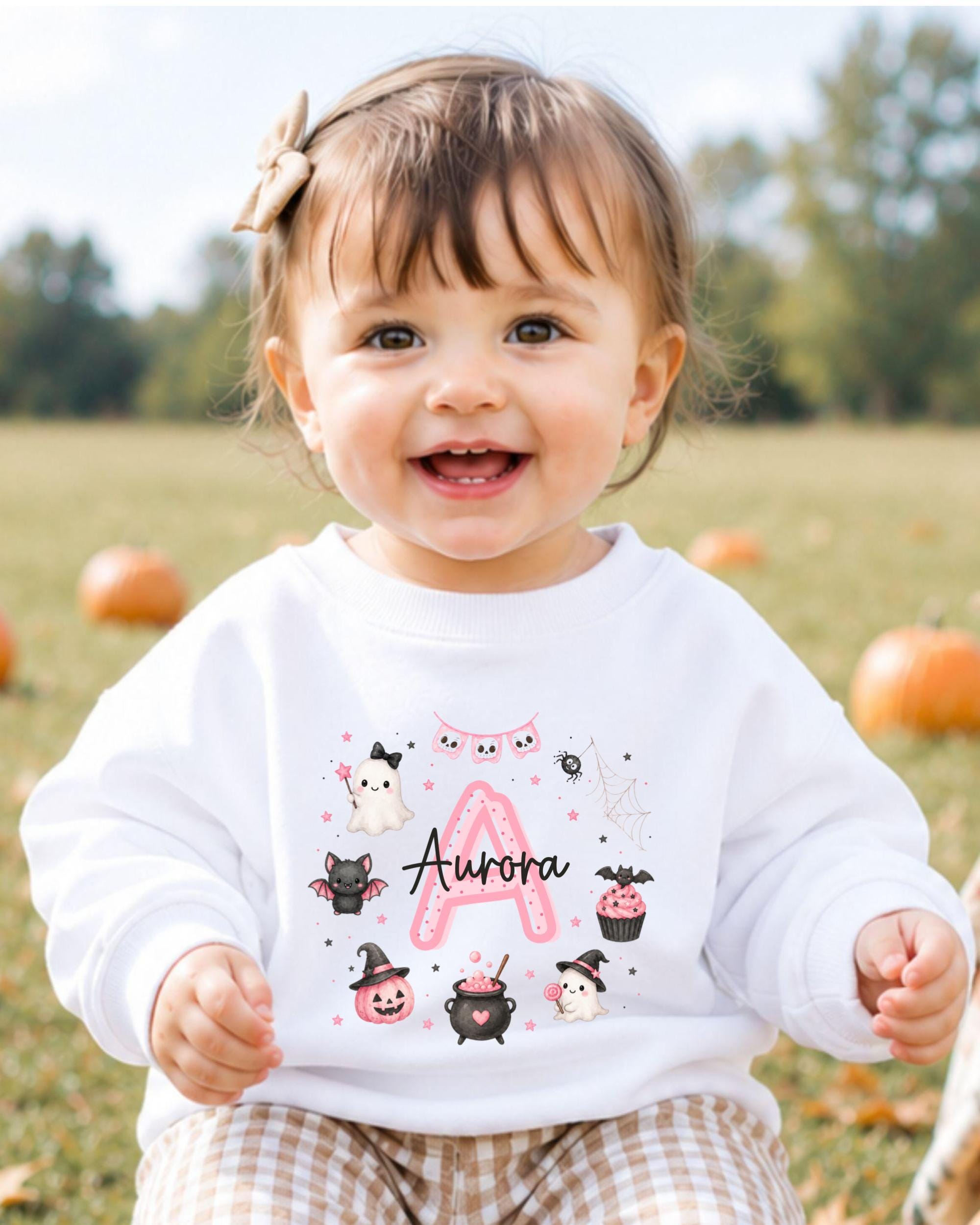 A smiling baby girl wearing a white sweater with a personalized name and Halloween-themed designs.